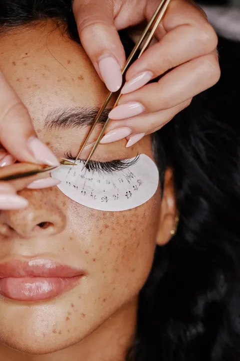 Eine Wimpernstylistin setzt mit zwei Pinzetten einzelne Wimpern; die Kundin trägt ein Pad unter dem Auge